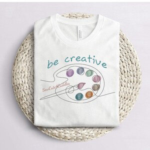 May include: White t-shirt with the words "be creative" in teal script above a line drawing of a paint palette filled with colorful yarn balls. The text "SewCuteCuddles" is below the palette.