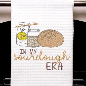 Puede incluir: Toalla de cocina blanca con tejido de gofres y diseño bordado. El diseño incluye una bolsa de harina, un tarro de granos y una hogaza de pan. El texto dice "IN MY sourdough ERA" en letras marrones y doradas.