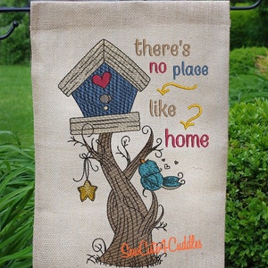 Può includere: Una bandiera da giardino in juta con una casetta per uccelli blu su un albero con un uccello blu e il testo "there's no place like home".