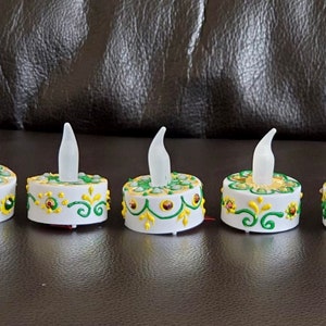 May include: Five white tea light holders with green and gold decorative designs. The holders have a small, clear, plastic flame in the center.
