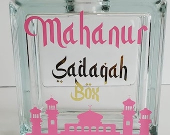May include: A glass block with a pink and gold design. The text "Mahanur Sadaqah Box" is written on the block. The block features a pink silhouette of a mosque.