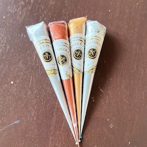 May include: Four henna cones with gold and white labels. The labels have the text "Imza Creations" and a logo. The cones are arranged in a fan shape with the tips pointing down.