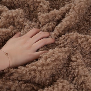 May include: Close-up of a soft, brown, fluffy fabric with a hand resting on it. The fabric has a curly, textured surface.