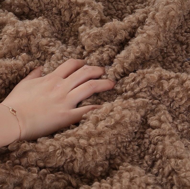 May include: Close-up of a soft, brown, fluffy fabric with a hand resting on it. The fabric has a curly, textured surface.