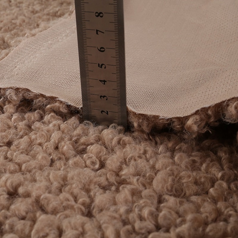 May include: Close-up of a beige faux fur fabric with a ruler measuring 8 inches. The fabric has a curly texture.