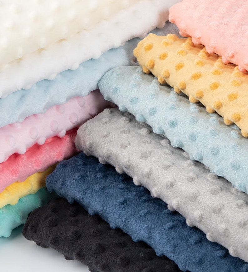May include: A stack of various colours of soft, textured fabric with a dotted pattern. The colours include white, light blue, pink, yellow, teal, grey, navy blue, and black.