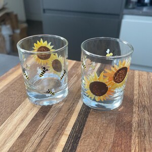 May include: Two clear glass shot glasses with painted sunflower and bee designs. The glasses are on a wooden surface.