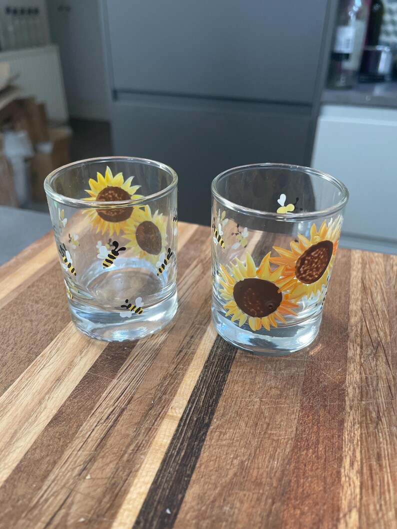 May include: Two clear glass shot glasses with painted sunflower and bee designs. The glasses are on a wooden surface.
