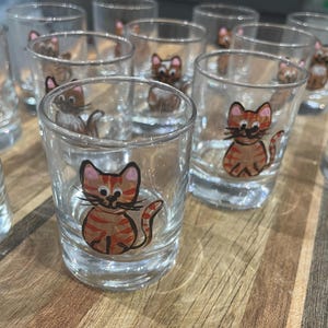 May include: Clear shot glasses decorated with hand-painted orange and brown tabby cat designs. Each glass features a smiling cat with striped markings. The glasses are displayed on a wooden surface.