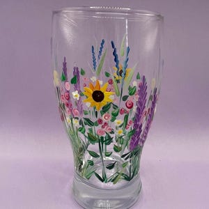 May include: A clear glass pint glass decorated with a hand-painted floral design. The artwork includes a yellow sunflower, purple lavender, pink roses, and other wildflowers. The glass is a standard pint glass shape.
