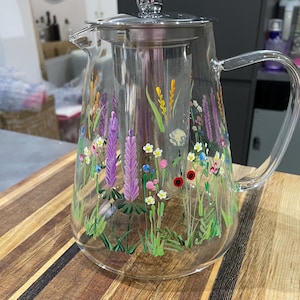 May include: A clear glass teapot with a stainless steel infuser and lid. The teapot is decorated with hand-painted wildflowers in shades of purple, yellow, red, and white. The teapot sits on a wooden surface.