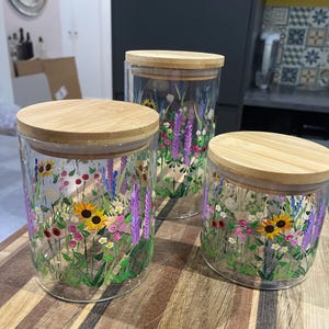 May include: Three clear glass storage canisters of varying heights, each topped with a round bamboo lid. The exterior of the glass is hand-painted with a colourful meadow scene featuring yellow sunflowers, purple spikes, and small white and pink flowers.