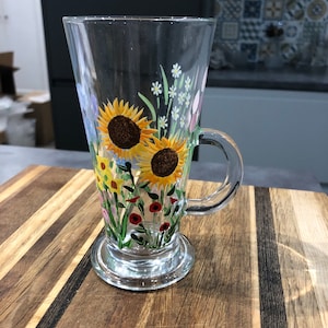 May include: A clear glass latte mug with a handle, hand-painted with sunflowers and wildflowers. The floral design includes yellow, orange, red, and white flowers with green stems and leaves. The mug sits on a clear, round base.