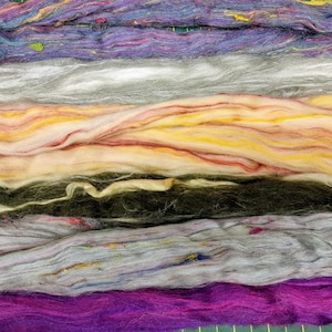 Könnte beinhalten: Nahaufnahme von mehreren Strängen farbiger Fasern. Die Fasern sind horizontal angeordnet und zeigen eine Vielzahl von Farben, darunter Lila, Grau, Gelb und Grün. Die Textur erscheint weich und flauschig, geeignet für Bastelarbeiten.