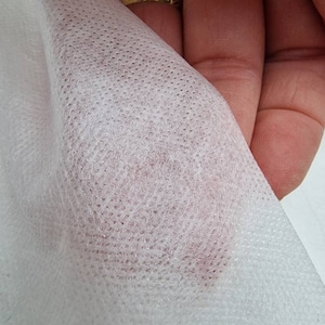 Może przedstawiać: Zbliżenie na białą, nie tkanej tkaninę o drobnej, kropkowanej fakturze. Tkanina jest lekko prześwitująca, ukazując delikatny różowy odcień pod spodem.