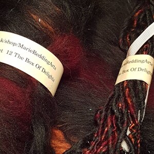 Op de afbeelding: Een bol zwart, rood en oranje garen met een wit label waarop staat "k/shop/Marie ReddingArts t 12 The Box Of Delights" en een ander wit label waarop staat "arie ReddingArts The Box Of Deligh"