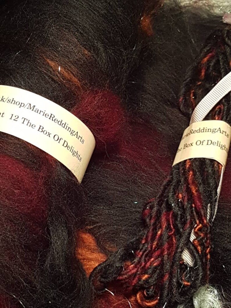 Op de afbeelding: Een bol zwart, rood en oranje garen met een wit label waarop staat "k/shop/Marie ReddingArts t 12 The Box Of Delights" en een ander wit label waarop staat "arie ReddingArts The Box Of Deligh"