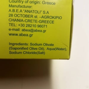 May include: A yellow box with black text listing the country of origin, manufacturer, address, phone number, email address, website, and ingredients for a product. The text reads: Country of origin: Greece Manufacturer: A.B.E.A "ANATOLI" S.A 28 OCTOBER st. -AGROK&Iota;&Rho;&Iota;&Omicron; CHANIA-CRETE-GREECE TEL: +30 28210 96071 e-mail: abea@abea.gr www.abea.gr Ingredients: Sodium Olivate (Saponified Olive Oil), Aqua (Water), Sodium Chloride(Salt)