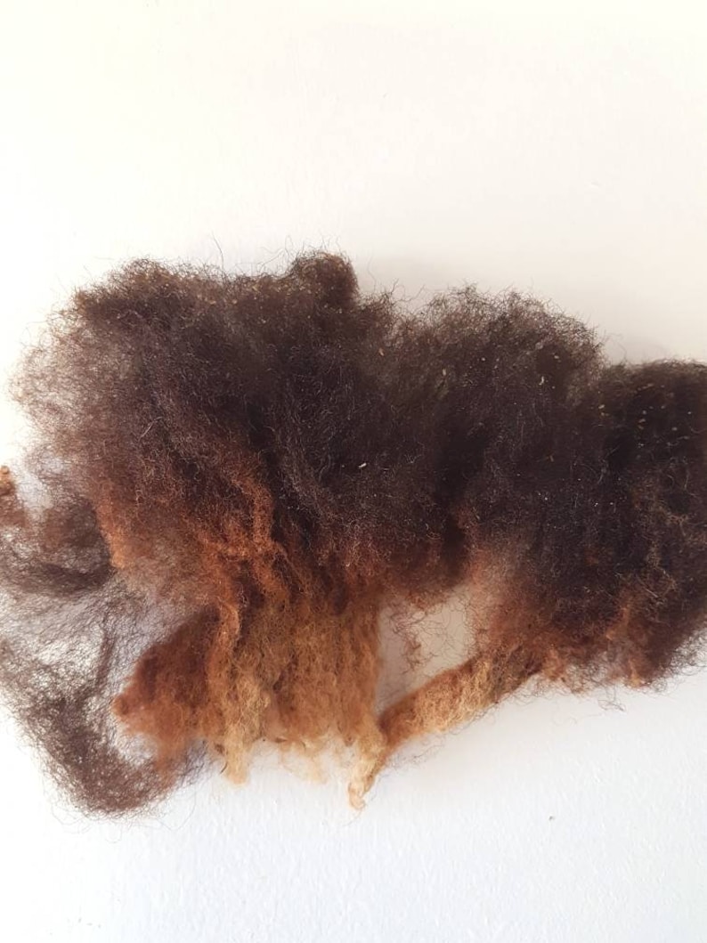 Op de afbeelding: Een close-up van een stapel donkerbruine en roodbruine wolvezels tegen een witte achtergrond. De wol heeft een pluizig, getextureerd uiterlijk, met verschillende tinten bruin en roodbruin.