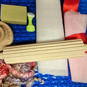 May include: A collection of craft supplies including a wooden rolling pin, a wooden stamp with text, a bar of soap, a green tool, pink fabric, red ribbon, and colorful wool fibers. The items are arranged on a blue textured surface.