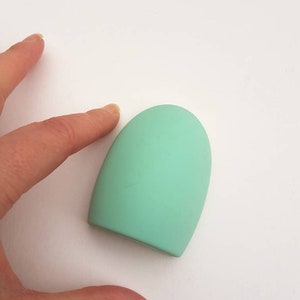 May include: A mint green, teardrop-shaped makeup brush cleaner. The silicone brush cleaner is a solid color and has a smooth, rounded top. The brush cleaner is shown on a white background.