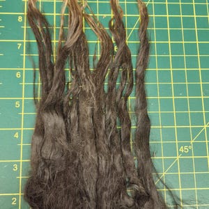 May include: A collection of long, dark brown hair strands with lighter, golden-brown tips, arranged vertically. The strands are placed against a green cutting mat with yellow grid lines, showing a length of approximately 20 cm.