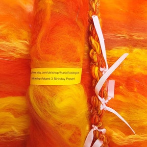 Puede incluir: Un rollo de lana naranja, amarilla y roja con una cinta rosa atada alrededor. La lana está etiquetada como "Fellowship Advent 3 Birthday Present".