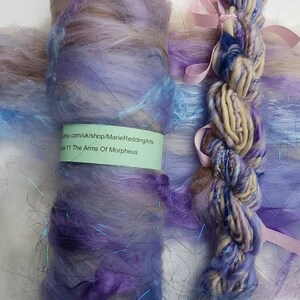 May include: A skein of hand-dyed yarn in shades of blue, purple, and beige with iridescent fibers. The yarn is labeled "The Arms of Morpheus" and is available for purchase at etsy.com/uk/shop/MarieReddingArts.