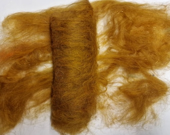 May include: A close-up of a golden brown wool roving, showing the fibers in a loose, fluffy state. The roving is partially rolled into a cylinder shape.