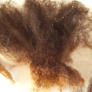 Op de afbeelding: Close-up van een stapel donkerbruine wolvezels. De vezels hebben een zacht, getextureerd uiterlijk met verschillende tinten bruin en bruin. Kleine vlekjes lichter materiaal zijn verspreid over de wol.