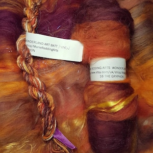 May include: A close-up of a multi-colored wool roving, with shades of orange, red, purple, and gold. The roving is arranged in a braid and a ball. The label reads "WONDERLAND ART BATT/YARN shop/MarieReddingArts HON" and "EREDDING ARTS WONDERLA www.etsy.com/uk/shop/Mar 16 THE GRYPHON".
