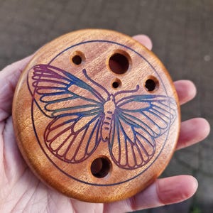 May include: A round wooden disc with a butterfly design engraved in the center. The butterfly is outlined in blue, green, and red. The disc has five holes drilled into it.