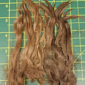 May include: Four distinct strands of brown hair fibers arranged vertically on a green cutting mat with a yellow grid. The strands vary in shade from light brown to dark brown, with a length of approximately 7 inches as indicated by the grid.