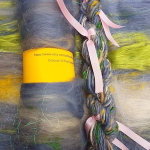 May include: A skein of blue, green and brown yarn with a yellow, green and blue fibre roving. The yarn is tied with pink ribbons and the roving is wrapped around a yellow tube with the text "https://www.etsy.com/uk/shop/Dracula13TheEscape" and "Dracula 13 The Escape".