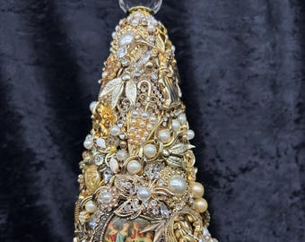 May include: A decorative gold Christmas tree sculpture adorned with an array of vintage jewelry, including pearls, rhinestones, and ornate metalwork. The tree features a clear, faceted crystal at the top and a small framed image on the side.