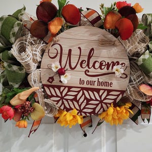 May include: A decorative wreath with a wooden sign that reads "Welcome to our home." The wreath features green and tan plaid ribbons, faux flowers, and fall-themed accents in shades of orange, red, and yellow.