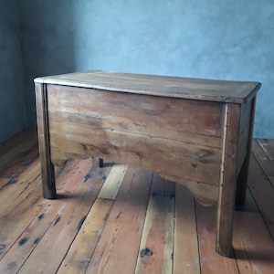 May include: A wooden chest with a light brown finish. The chest has a rectangular top and four legs. The chest is sitting on a wooden floor.