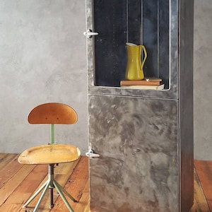 May include: A vintage metal cabinet with glass doors and a wood top. The cabinet has a distressed silver finish and is sitting on a wooden floor. A yellow pitcher and books are on the top shelf. A wooden chair with a green metal base is in the foreground.