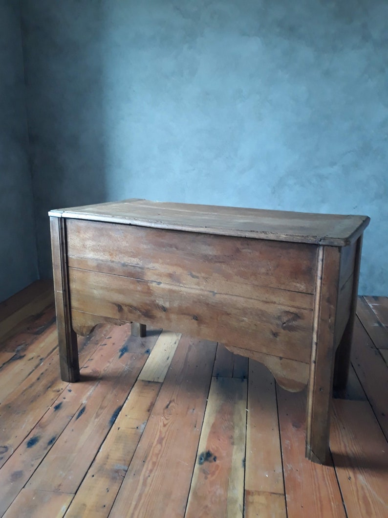 May include: A wooden chest with a light brown finish. The chest has a rectangular top and four legs. The chest is sitting on a wooden floor.