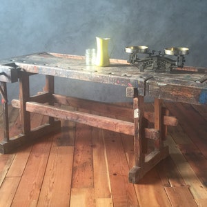 May include: A vintage wooden workbench with a weathered, distressed finish. The workbench has a vise on the left side and a metal scale on the top. The workbench is sitting on a wooden floor.