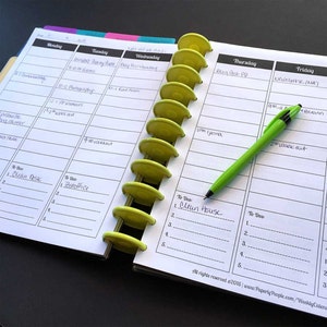 May include: Open weekly planner with a green pen, and yellow disc-shaped dividers. The planner pages are white with black lines and text, including daily schedules and to-do lists. The text includes the phrase "Clean Desk".