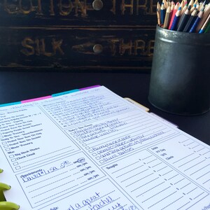 May include: A close-up of a white planner with blue ink. The planner has a tabbed section for each day of the week. The planner is open to Wednesday, August 16th. The planner has a section for appointments, to-do list, and notes. The planner is sitting on a black surface with a black tin filled with colored pencils.