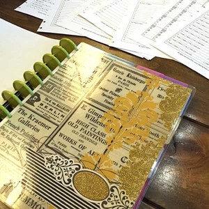 May include: A black and white planner with a gold glitter cover. The cover features vintage-style advertisements for art galleries in Paris and Berlin. The planner has a green binder with silver rings.