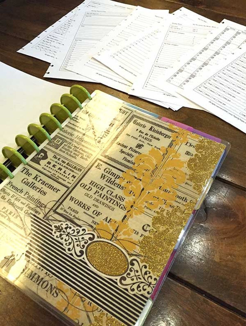 May include: A black and white planner with a gold glitter cover. The cover features vintage-style advertisements for art galleries in Paris and Berlin. The planner has a green binder with silver rings.