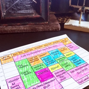 May include: A colorful weekly planner with handwritten notes and highlighted sections. The planner includes tasks like "Blog Post," "Client Work," and "Kids Home/Homework." The planner is on a dark surface.
