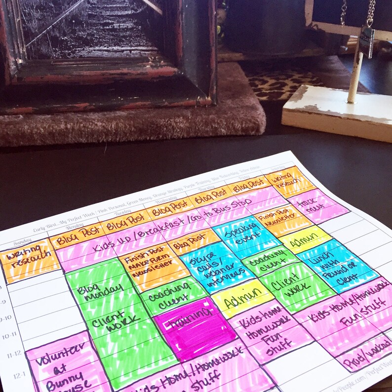 May include: A colorful weekly planner with handwritten notes and highlighted sections. The planner includes tasks like "Blog Post," "Client Work," and "Kids Home/Homework." The planner is on a dark surface.