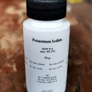 Może przedstawiać: Biała plastikowa butelka z czarną zakrętką z etykietą "Potassium Iodide" z informacjami o czystości, wadze i składzie chemicznym. Etykieta zawiera również datę 20/12.2025.