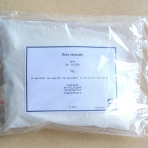 Può includere: Un sacchetto di plastica trasparente contenente una polvere bianca. Un'etichetta bianca sulla busta riporta "Zinc stearate" con informazioni chimiche, tra cui "Zn - 10-12%" e "50g."