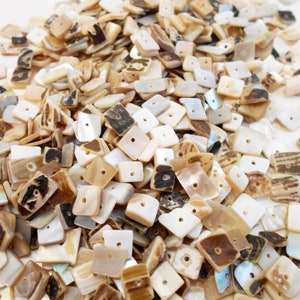 May include: A large pile of small, square, iridescent shell beads with a hole in the center. The beads are a mix of white, brown, and beige.