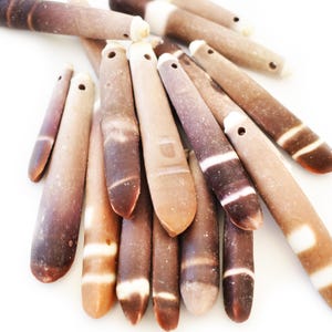 May include: A collection of elongated, cylindrical beads in varying shades of brown, tan, and white. Each bead has a hole drilled through one end. The beads have a natural, organic appearance, with striped and mottled patterns.